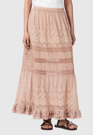 Kvinde iført en lyserød broderet nederdel til anklerne med flæsekant og sorte sandaler på en ensfarvet baggrund.