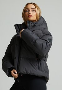 Giacca a vento imbottita di colore grigio scuro con colletto alto, chiusura con zip frontale e tasche laterali. Presenta dettagli neri e una texture imbottita per l'isolamento.