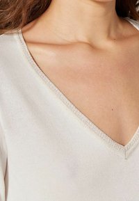 Bluzka z dekoltem w serek w jasnym kremowym materiale o gładkiej fakturze i subtelnym błysku wzdłuż linii dekoltu. Zbliżenie na dekolt i ramię.