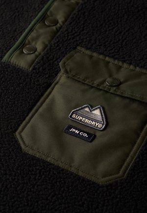 Sudadera negra de lana con un bolsillo de nylon verde oliva con botones a presión, logo bordado y botones a juego. Textura suave con un contraste de color distintivo.