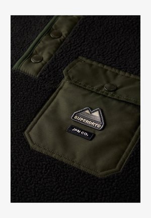 Sort fleece-pullover med en olivengrøn nylon snap-lomme, broderet logo og matchende knapper. Blød tekstur med tydelig farvekontrast.