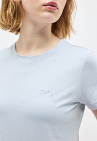 Νεαρή γυναίκα με κοντά ξανθά μαλλιά που φοράει ανοιχτό μπλε μπλουζάκι με το «BOSS» κεντημένο στο στήθος.