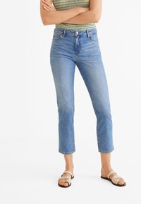 Mujer vestida con jeans cortos de talle alto en azul, sandalias beige y una blusa sin mangas de punto a rayas con los brazos cruzados.