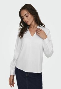 Blusa bianca a maniche lunghe con scollatura a V, caratterizzata da una consistenza liscia e maniche arricciate. Abbinata a una gonna in denim scuro.