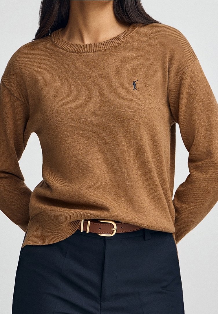 Femme portant un pull marron à col rond avec un petit logo de golfeur brodé, rentré dans un pantalon noir fixé par une ceinture en cuir marron.
