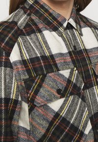 Camicia di flanella con motivo a quadri nei toni del nero, rosso, bianco e giallo. Include bottoni e due tasche sul petto.