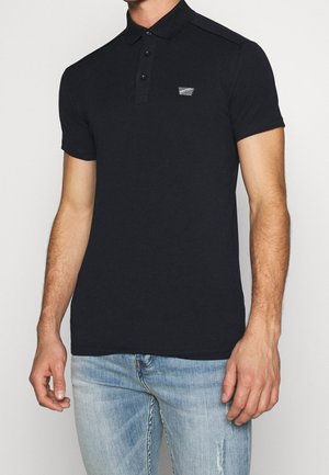 Homme portant un polo noir cintré à manches courtes avec trois boutons et un petit logo rectangulaire sur la poitrine, associé à un jean bleu clair.