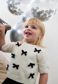 Lächelndes Kind mit blonden Haaren, das einen weißen Pullover mit schwarzen Schleifen trägt und ein Geländer hält, im Hintergrund silberne und durchsichtige Ballons.