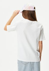 Vit bomulls t-shirt med kort ärm, rund halsringning och avslappnad passform. Bärs med en pastellfärgad tie-dye keps och ljusblå denimkjol.