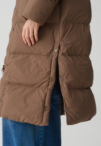 Brauner Puffer-Mantel mit gestepptem Design, ausgestattet mit Druckknöpfen und einem leichten Schlitz am Saum. Die Materialien wirken leicht und texturiert.