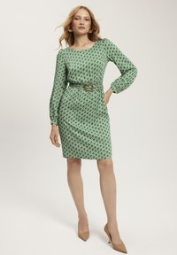 Zielona sukienka w geometryczny wzór z długimi rękawami, dopasowaną sylwetką, okrągłym dekoltem i brązowym paskiem. Noszona z beżowymi szpilkami.