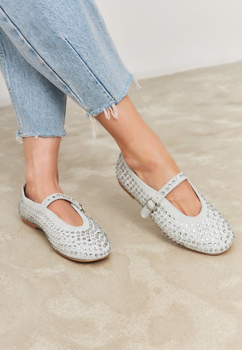 Stopy w jasnoniebieskich dżinsach z postrzępionymi brzegami i jasnoszarej baletowej obuwie Mary Jane ozdobione okrągłymi srebrnymi ćwiekami na beżowej podłodze.