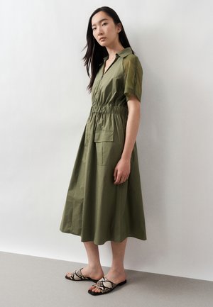 Kvinde står op ad hvid væg iført olivengrøn midi-kjole med korte ærmer, elastik i taljen og lomme, kombineret med sandaler med slangeprint.