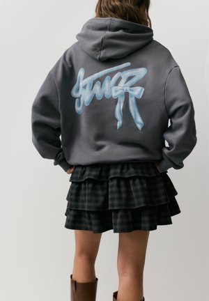 Osoba v šedé mikině s kapucí s modrým graffiti textem na zádech, vrstvenou tmavou károvanou sukní a hnědými kozačkami po kolena, stojící s rukama v bok.