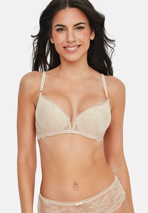 Femme souriante aux longs cheveux noirs portant un soutien-gorge en dentelle crème et une culotte assortie, sur un fond uni.