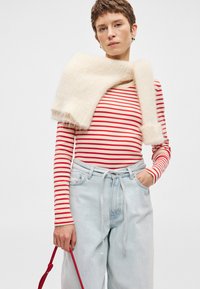 Ριγέ μπλούζα με μακριά μανίκια σε κόκκινο και μπεζ χρώμα, συνδυασμένη με ανοιχτό μπλε φαρδύ παντελόνι. Ένα απαλό κρεμ κασκόλ κρέμεται στους ώμους. Λεπτομέρεια κόκκινης τσάντας.