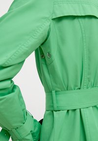 Gros plan sur une veste verte montrant une taille ceinturée, une manche avec une sangle et des œillets de ventilation sur un tissu lisse.