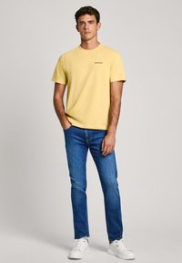 T-shirt en coton jaune avec un col rond et des manches courtes, associé à un jean en denim bleu et des baskets blanches. Look simple et décontracté.
