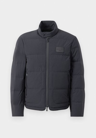 Doudoune noire avec un col montant, texture matelassée et fermeture éclair à l'avant. Elle possède une petite étiquette avec logo sur la poitrine gauche et des poches latérales.