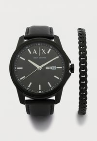 Črna silikonska ura z okroglo številčnico, belimi oznakami ur, prikazom datuma in črnim usnjenim pasom. Vključuje črno verižico.