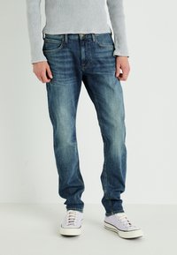 Jasnoniebieskie dżinsy w prostym kroju, z delikatnym wyblaknięciem i standardowym designem pięciu kieszeni. Noszone z białymi i lawendowymi sneakersami.