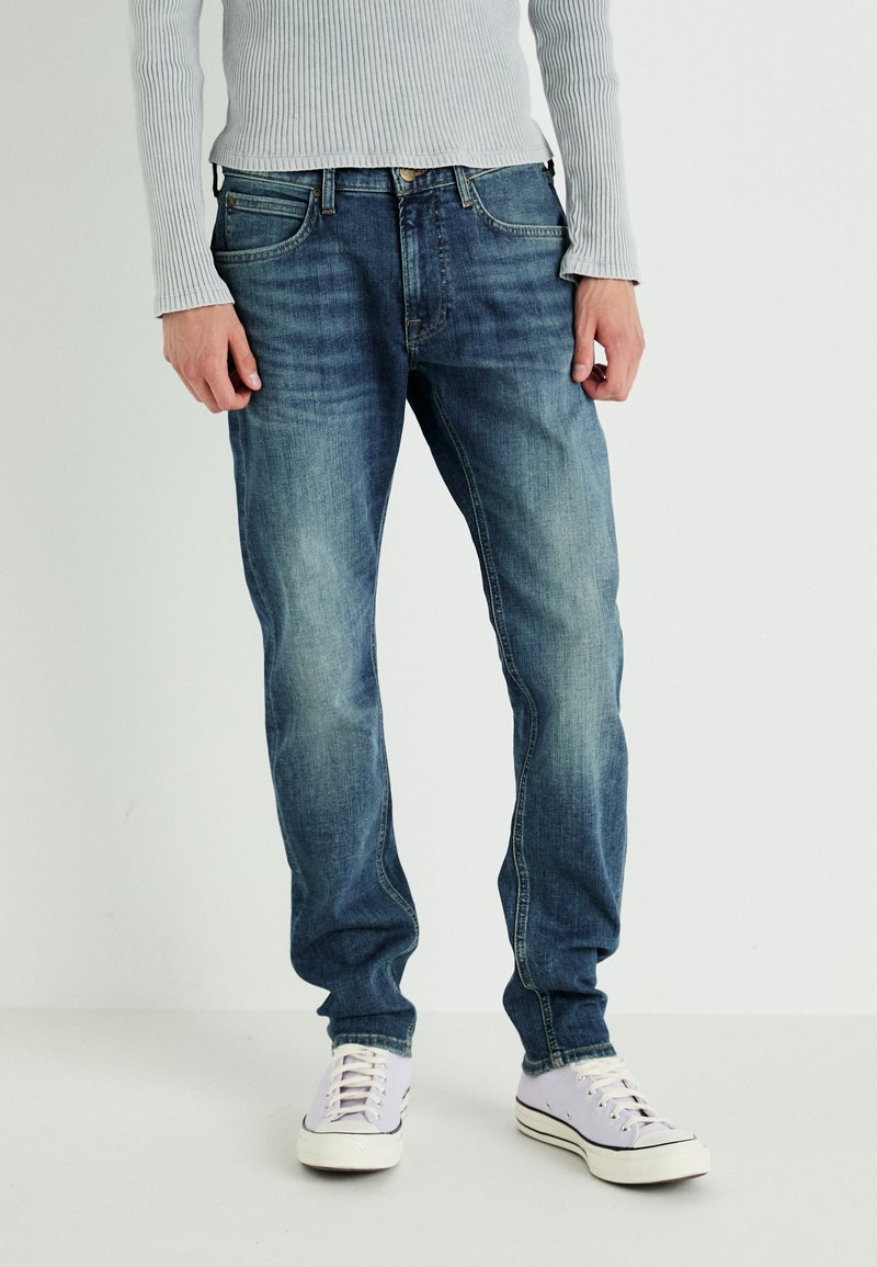 Jasnoniebieskie dżinsy w prostym kroju, z delikatnym wyblaknięciem i standardowym designem pięciu kieszeni. Noszone z białymi i lawendowymi sneakersami.