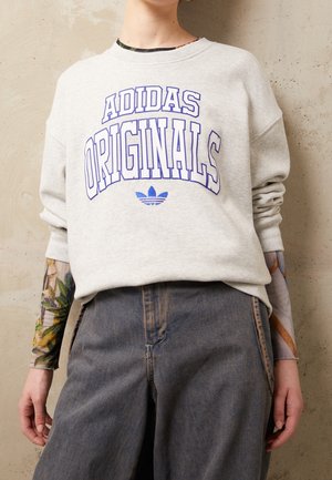 Personne portant un sweat-shirt Adidas Originals gris clair avec un logo bleu, un pantalon ample gris foncé et une chemise à manches longues à motif floral en dessous.