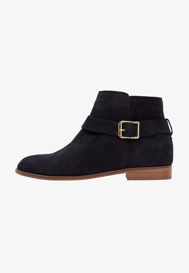 Bottine en suède noir avec une boucle latérale dorée, petit talon en bois et bout arrondi, présentée en profil sur un fond blanc.