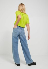 Kvinna med axellångt blont hår som bär en limegrön t-shirt, högmidjade vida blå jeans och svarta kraftiga skor, står med ryggen vänd.