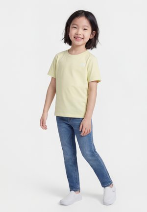 Lächelnes Mädchen, das ein hellgelbes kurzärmeliges Shirt, blaue Jeans und weiße Slipper trägt, steht vor weißem Hintergrund.