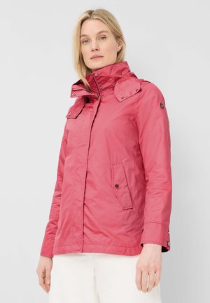 Frau mit blondem Haar, die eine rote Jacke mit hohem Kragen und Knöpfen sowie weiße Hose trägt, vor einem hellgrauen Hintergrund stehend.