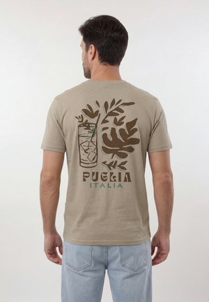 Férfi, aki bézs pólót visel barna és zöld növényi mintával, kezében egy pohár jeges ital, a póló hátán a „Puglia Italia” felirat.