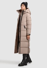 Hosszú, bélelt kabát világos bézs színben, kapucnival, cipzáras zsebekkel és fekete részletekkel. Fekete fűzős csizmával és sapkával párosítva.