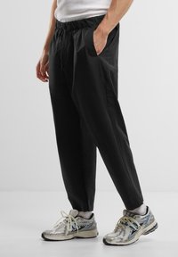 Pantaloni negri, largi, cu talie elastică, buzunare laterale și o textură netedă, asortați cu adidași argintii.
