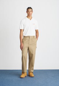 Polo bianco con logo arancione, pantaloni beige e stivali da lavoro gialli. Il modello si trova davanti a uno sfondo semplice con pavimentazione blu.