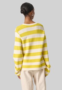 Pull rayé en jaune et crème, avec une coupe décontractée, une texture côtelée, des manches larges et un ourlet arrière légèrement raccourci. Toujours élégant.