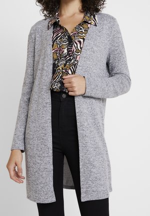Cardigan grigio con scollo aperto e maniche lunghe, realizzato in un tessuto testurizzato. Indossato sopra una camicia a fantasia con sfondo nero che presenta design colorati.