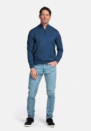 Man die recht naar voren kijkt, gekleed in een blauwe trui met een kwart-ritssluiting, lichtblauwe spijkerbroek en zwarte sneakers, met één hand in de zak.