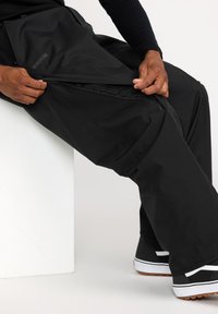 Pantalons imperméables noirs avec une ouverture zippée, présentant un tissu texturé. Accentué d'un logo et associé à des baskets noir et blanc.