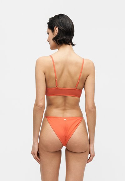 Γυναίκα με κοντά σκούρα μαλλιά φορώντας πορτοκαλί μπικίνι με ρυθμιζόμενα λουριά, στέκεται με την πλάτη προς την κάμερα σε απλό φόντο.