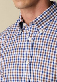 Chemise à carreaux en bleu, marron et blanc avec un col boutonné, ornée de petits carrés et de boutons standards le long de la patte de boutonnage.
