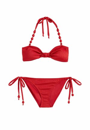 Žena v červenom bikiny a slnečných okuliaroch kráča bosá pozdĺž pobrežia pláže, nesúc pletenú tašku, v pozadí palmy a oceán.