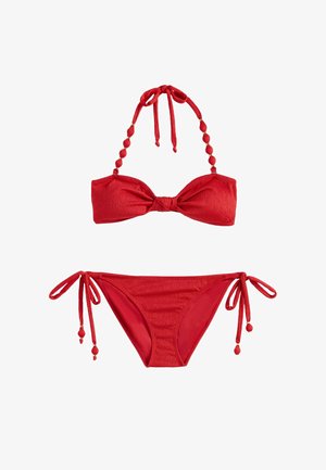 Femme en bikini rouge et lunettes de soleil marchant pieds nus au bord de la plage, portant un sac tressé, palmiers et océan en arrière-plan.