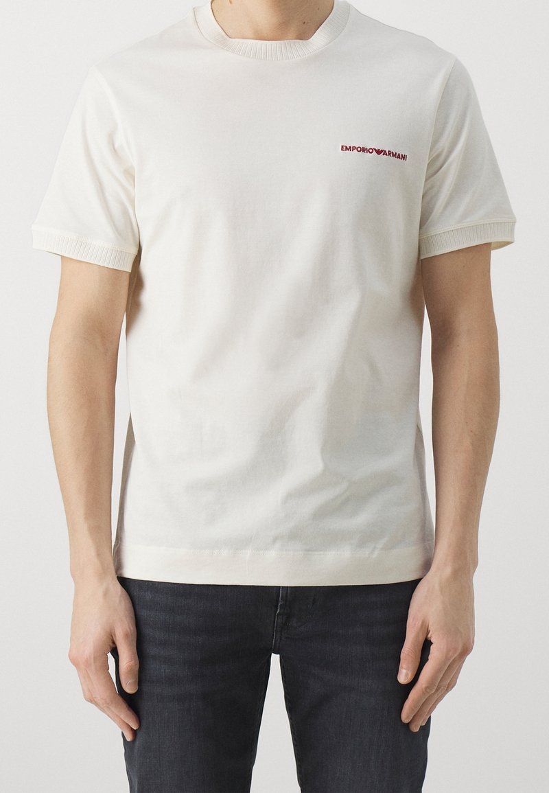 Biała bawełniana koszulka z krótkim rękawem, prążkowanym dekoltem i subtelnym czerwonym logo "EMPORIO ARMANI" na lewej piersi. Prosta, gładka tekstura.