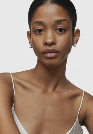 Orecchini con un design geometrico caratterizzati da metallo argenteo e accenti di pietre trasparenti. Indossati da una modella in un top bianco a spalline sottili.