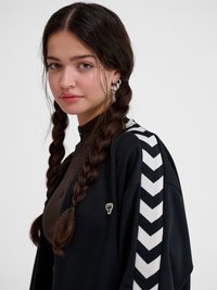 Młoda kobieta z długimi splecionymi włosami nosi czarną kurtkę z białymi paskami w kształcie jodełki i srebrne kolczyki koła, patrzy lekko na bok.