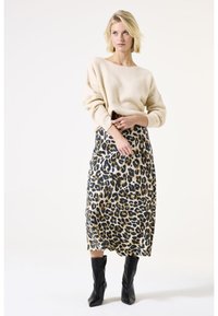 Beige gestrickter Pullover, midi Rock mit Leopardenmuster in Beige, Schwarz und Braun, kombiniert mit schwarzen Stiefeletten. Model posiert mit den Händen in den Hüften.