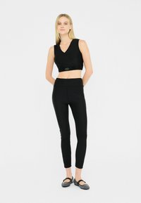 Top corto nero con scollo a V e vita elasticizzata, abbinato a leggings neri a vita alta. Il modello indossa delle ballerine nere. Tessuto liscio.