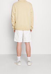 Beige sweatshirt med krage, långa ärmar och ribbade muddar; kombinerad med vita shorts och grå sneakers. Enkel design med ledig passform.