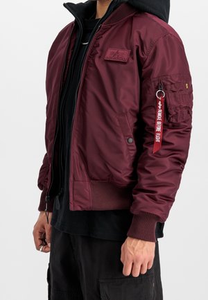 Homme portant une veste bomber bordeaux avec des poignets et un ourlet côtelés, une étiquette "Remove Before Flight" sur la poche de la manche gauche, et des vêtements noirs en dessous.